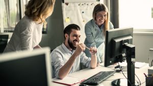 3 Kolleg:innen stehen im Büro vor einem Monitor und lachen